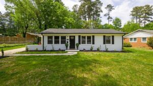 Classic single-story white ranch-style home with black shutters, surrounded by green trees and a wide front lawn.