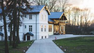 Charming white two-story traditional cottage with a dark metal roof, brick chimney, and surrounding trees.
