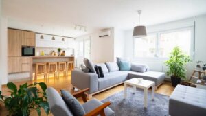Modern open-concept living room with a large grey sectional sofa, wooden floors, and a kitchen background.