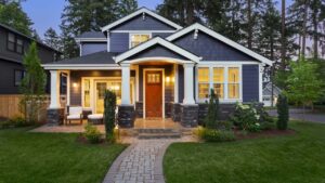 Craftsman-style two-story blue house with stone pillars, a warm front porch light, and detailed gables.